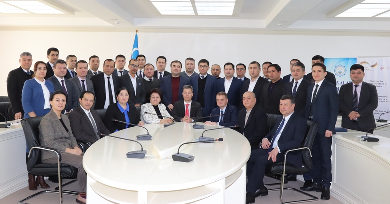 Ein Gruppebild zeigt viele Männer und einige Frauen in Anzügen, die im Halbkreis um einen runden Tisch im usbekischen Verwaltungsrichter angeordnet sind.