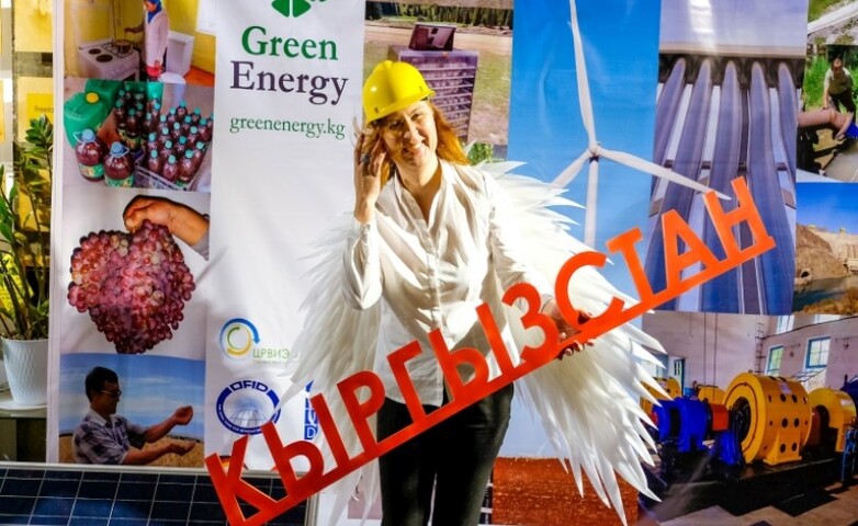 Eine junge Frau steht vor einem Plakat für eine Handelsmesse zum Thema umweltfreundliche Energie und hält einen Gegenstand in der Hand, der an einen Schriftzug erinnert.