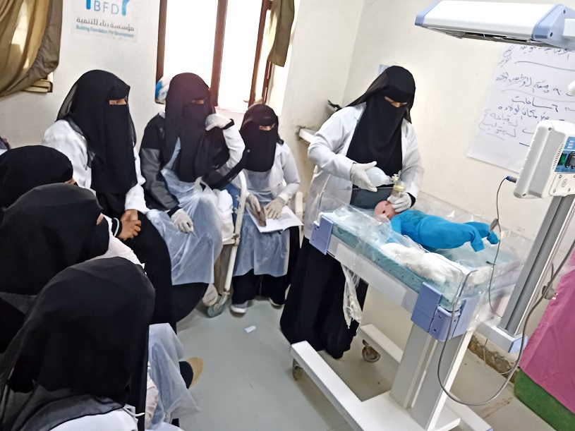 Eine Gruppe von Frauen beobachtet eine Frau, die medizinische Handgriffe an einer Babypuppe vornimmt. 