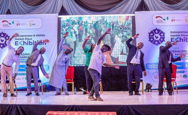 Bei einer Konferenz zu Unternehmertum stehen sieben Personen auf einer Bühne und heben lächelnd die Arme in die Luft.