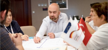  Ein Mann zeichnet etwas auf einem Dokument ein. Um ihn herum sitzen weitere Personen und auf dem Tisch stehen kleine Flaggen, darunter die der EU und die der Türkei.