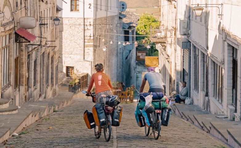 Two people are riding down a cobblestone street on heavily laden bicycles.