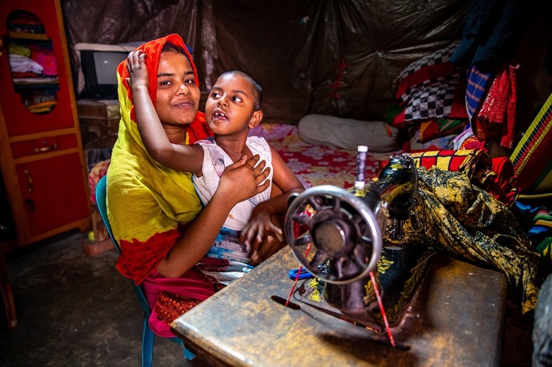 A self-employed seamstress sits at a sewing machine with her son. 