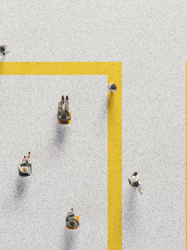 Luftaufnahme von Menschen, die in einer hellen Umgebung und in gleichmäßigen Abständen auf Stühlen sitzen oder über gelb markierte Wege laufen.