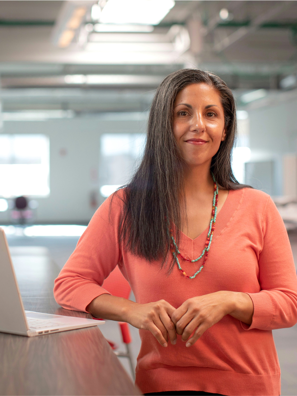 Eine Frau steht lächelnd an einem Tisch in einem Bürogebäude. Neben ihr steht ein aufgeklappter Laptop.  