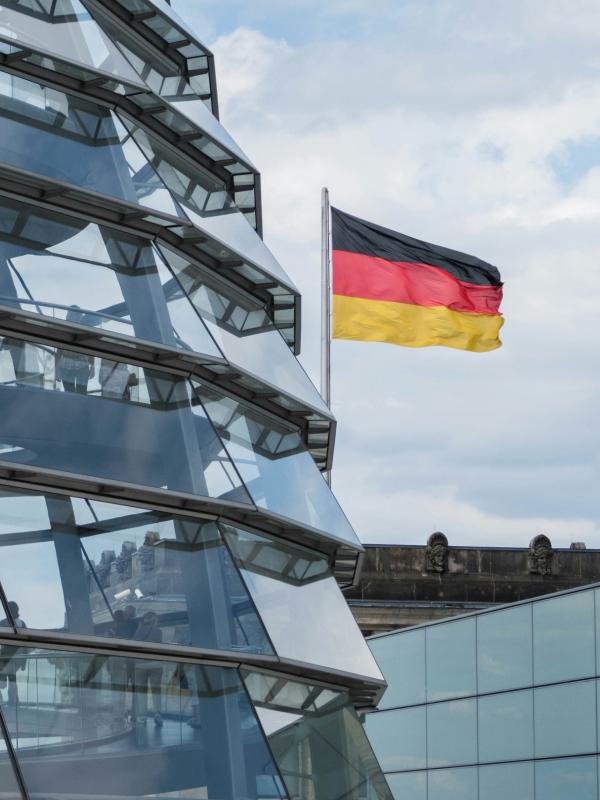 Ausschnitt der gläsernen Bundestagskuppel vor einer Deutschlandfahne und bewölktem Himmel. In der Kuppel sind viele Menschen zu sehen.