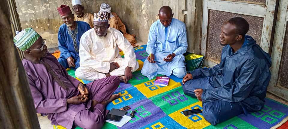 A group of men sit on a colorful mat, engaged in discussion