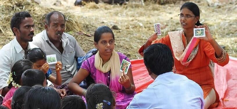 A group of people sit outside on a red tarpaulin and play cards.