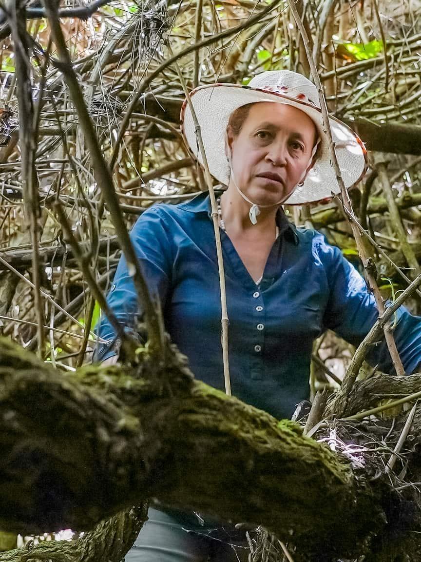 Eine Frau mit breitkrempigem Hut und blauem Hemd steht inmitten dichter, ineinander verwobener Äste in einer Waldlandschaft.