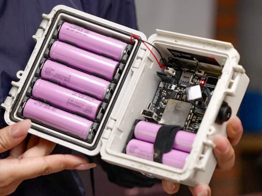 Eine Person hält ein offenes Gerät mit sechs pinken zylindrischen Batterien und sichtbaren elektronischen Komponenten in einem Kunststoffgehäuse.