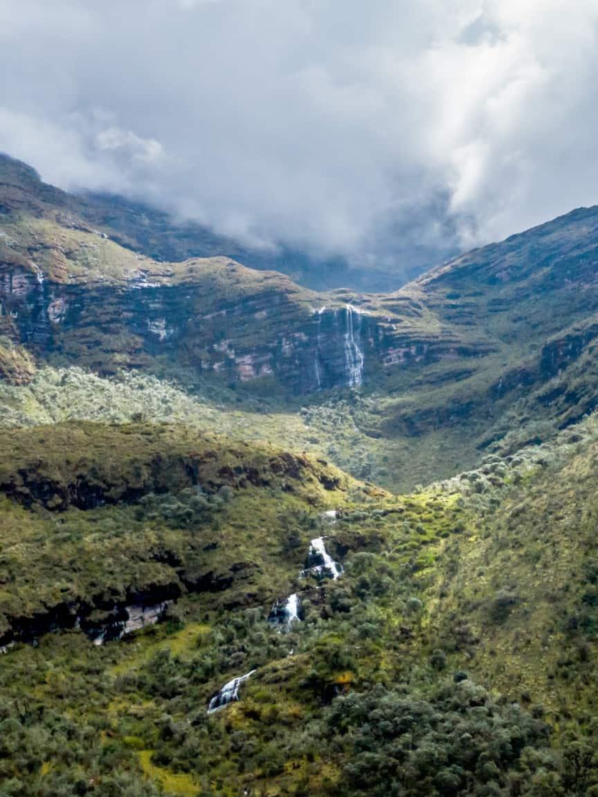 Eine malerische Aussicht auf ein grünes Bergtal mit mehreren Wasserfällen, die herabfließen, teils von Wolken bedeckt.