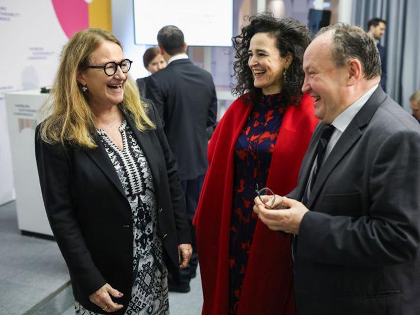 Three people in business attire laugh and chat at an event; one woman wears a black and white dress, another wears a red coat, and one man wears a suit.