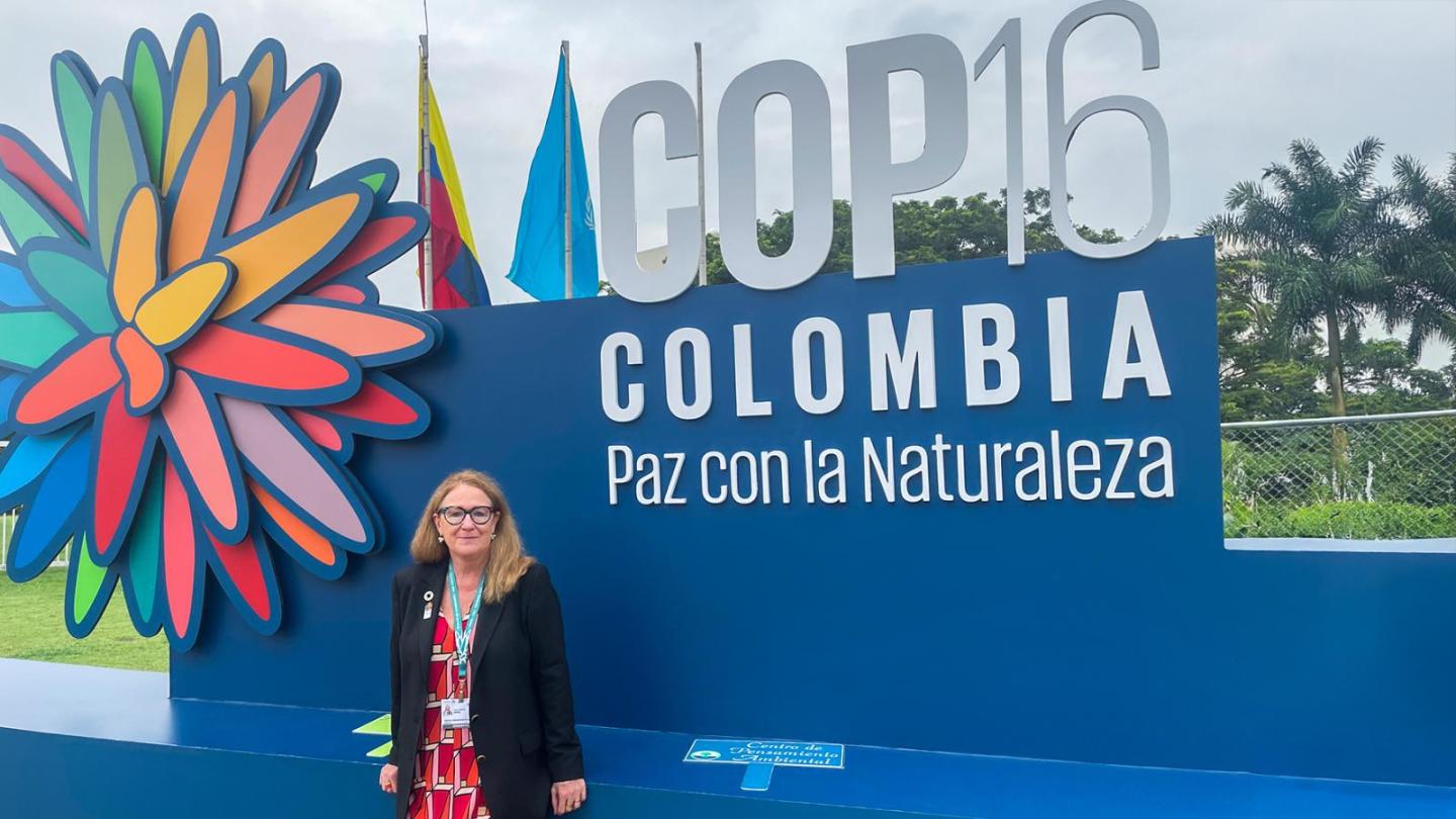 A woman stands in front of a sign reading ‘COP16 Colombia Paz con la Naturaleza’ outdoors, wearing a black jacket and a patterned dress.
