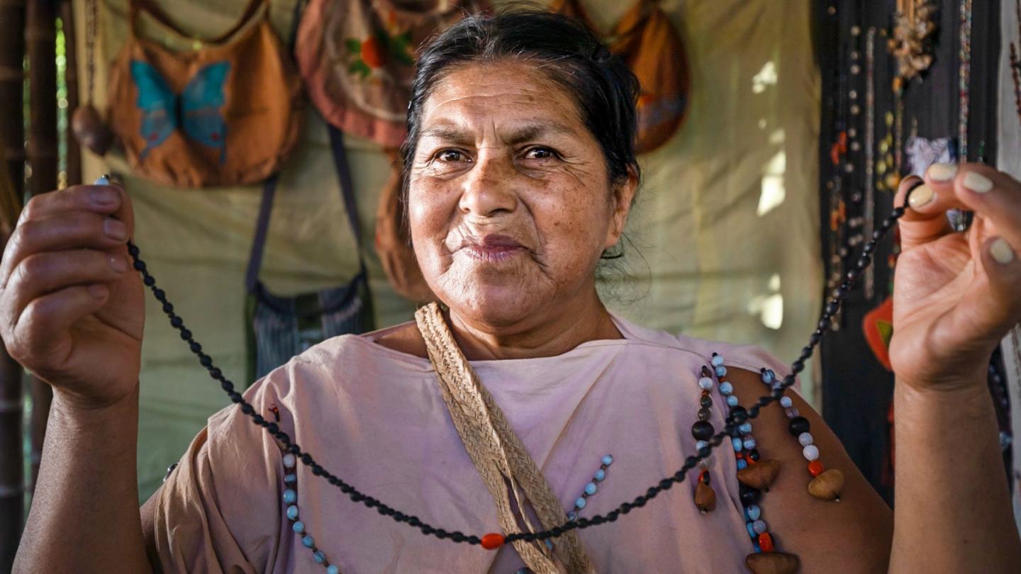 An indigenous woman holds a handmade necklace in front of the camera.