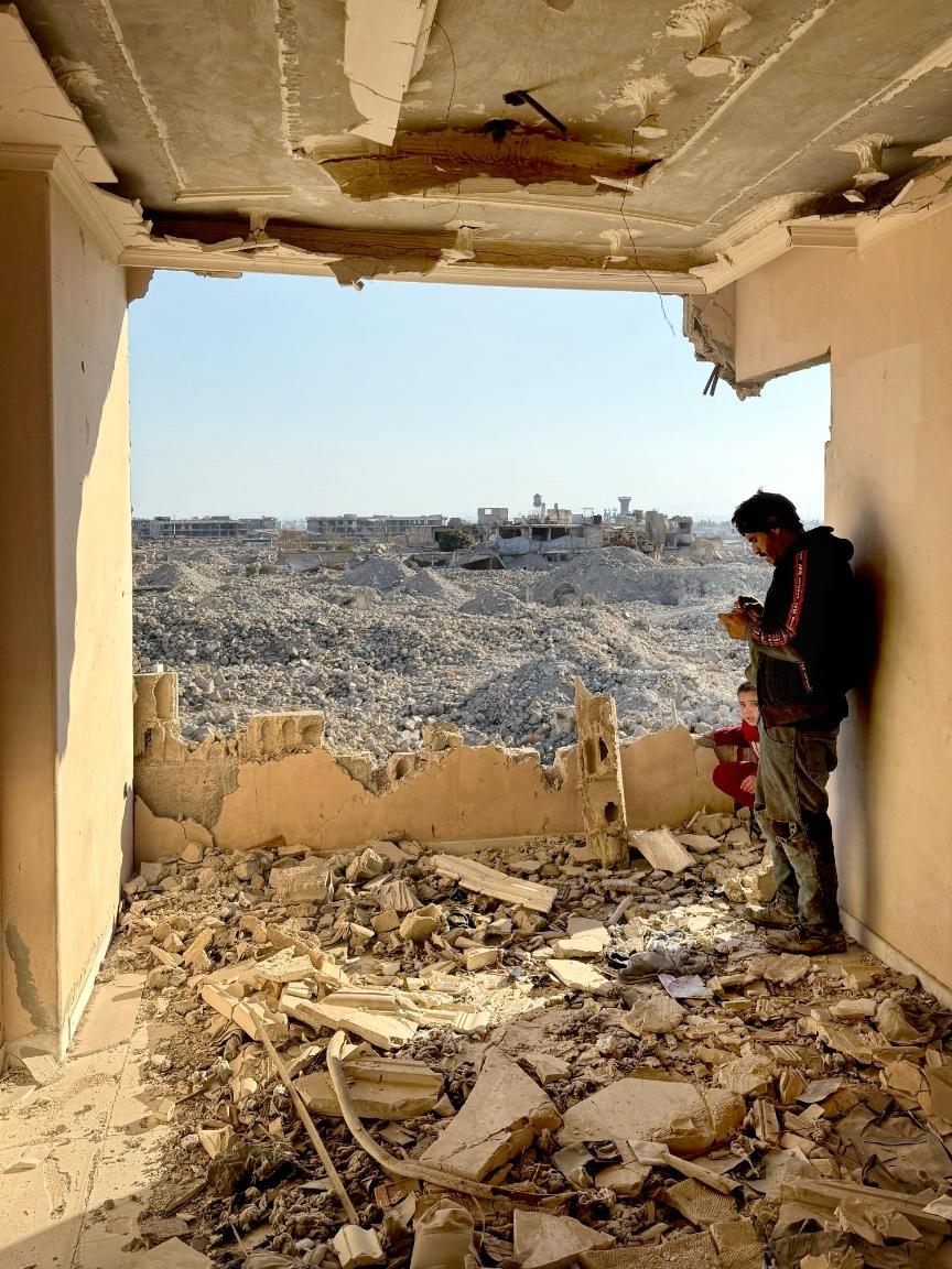 View from a destroyed building with a sagging ceiling onto a vast field of rubble, with a man standing in the foreground and a child in the corner.