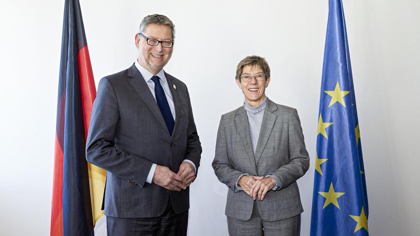 Two people in formal attire stand side by side and smile at the camera, flanked by a German flag on the left and a European flag on the right against a light background.