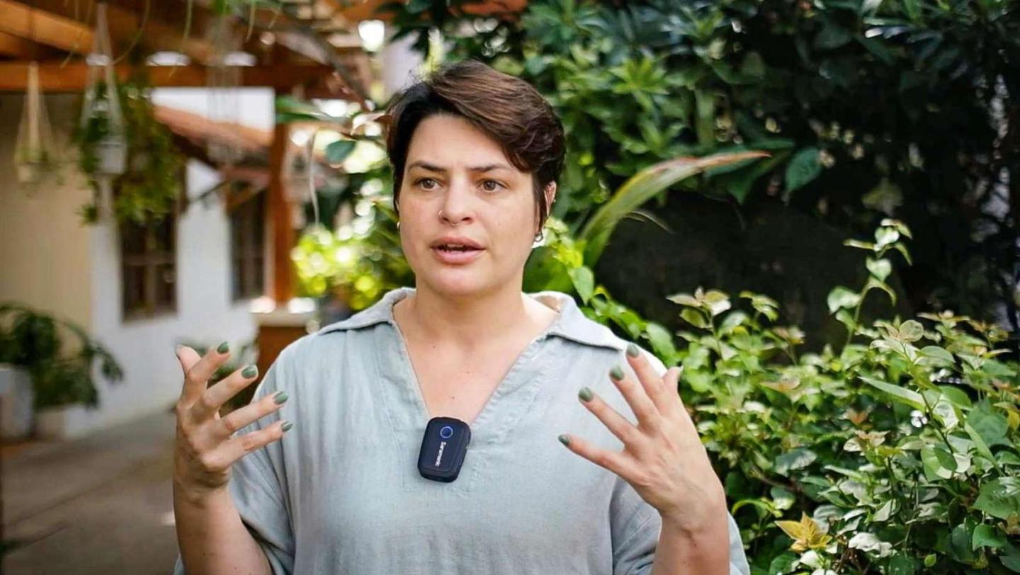 Eine Frau mit Kurzhaarschnitt spricht gestikulierend vor tropischem Grün und trägt ein Mikrofon am Hemd.