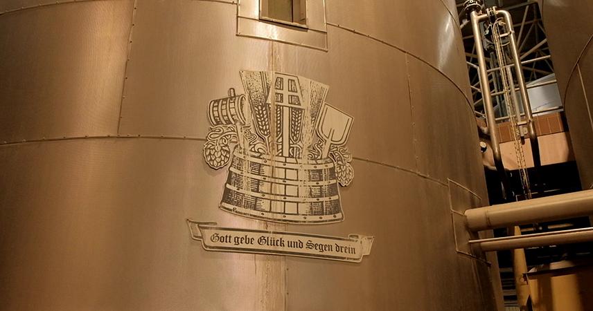 An engraved emblem on a wooden surface depicting traditional brewery symbols, with a beer crown in the centre, flanked by ears of barley and possibly brewing tools. Below the emblem is a banner with the inscription “God grant good fortune and blessings”.