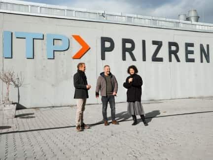 Drei Personen stehen vor einer grauen Wand mit der Aufschrift &quot;ITP PRIZREN&quot; und unterhalten sich.
