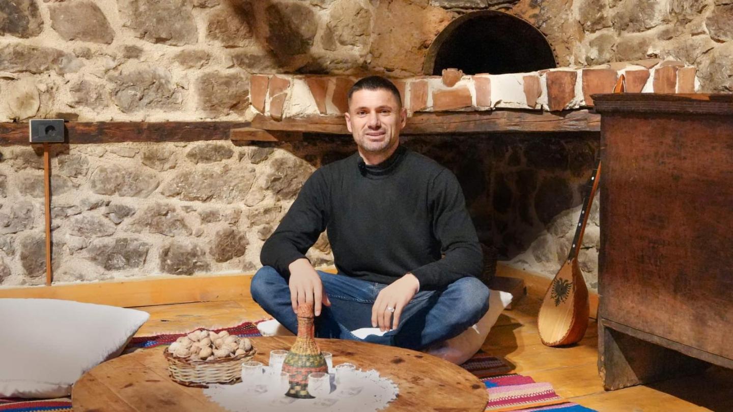 Ein Mann in schwarzem Pullover und Jeans sitzt auf dem Boden in einem gemütlichen Raum mit Steinmauern und einem alten Backofen.