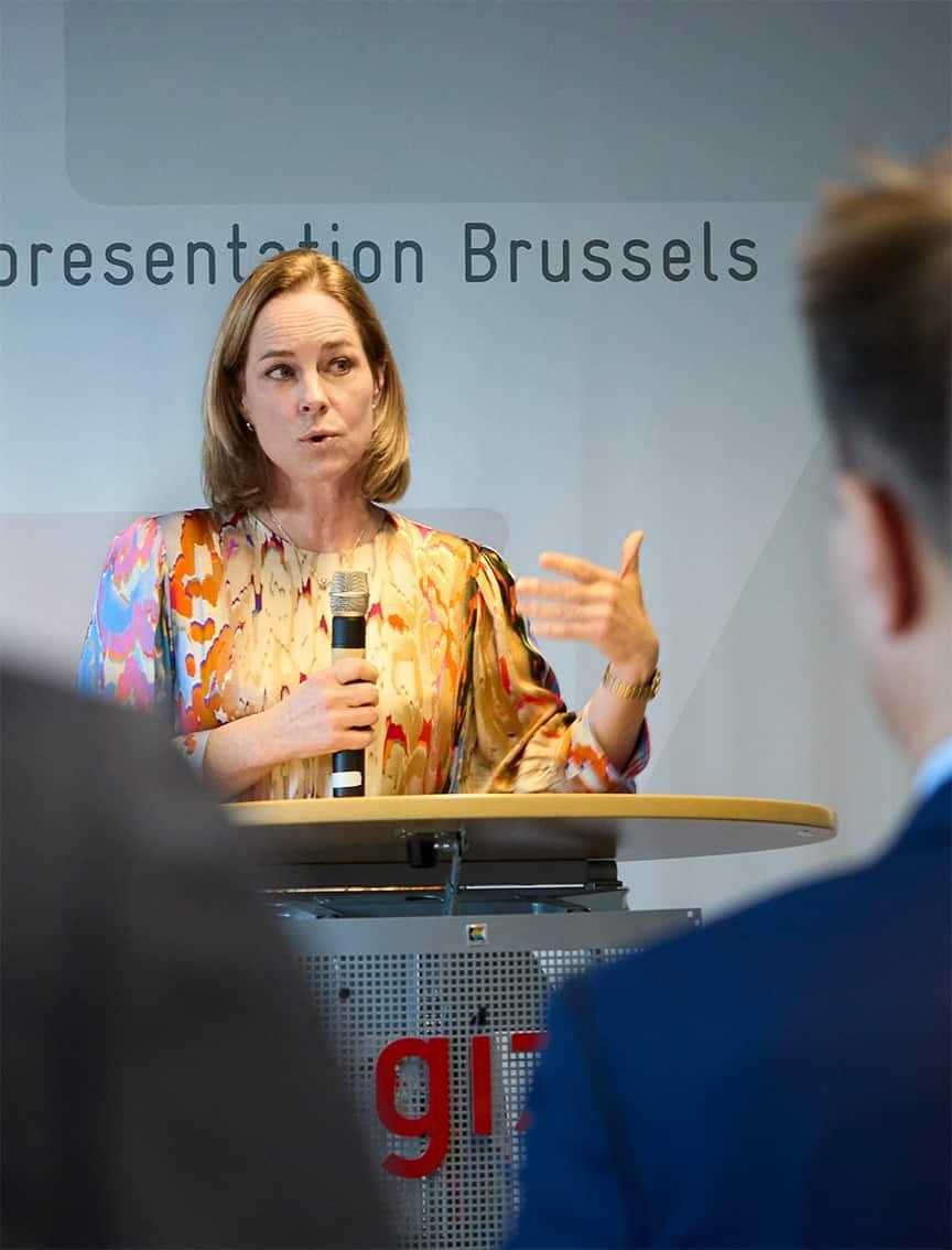 Eine Frau mit schulterlangem blondem Haar trägt eine farbenfrohe Bluse mit abstraktem Muster und spricht auf einer Veranstaltung in Brüssel an einem Rednerpult mit einem Mikrofon in der Hand.