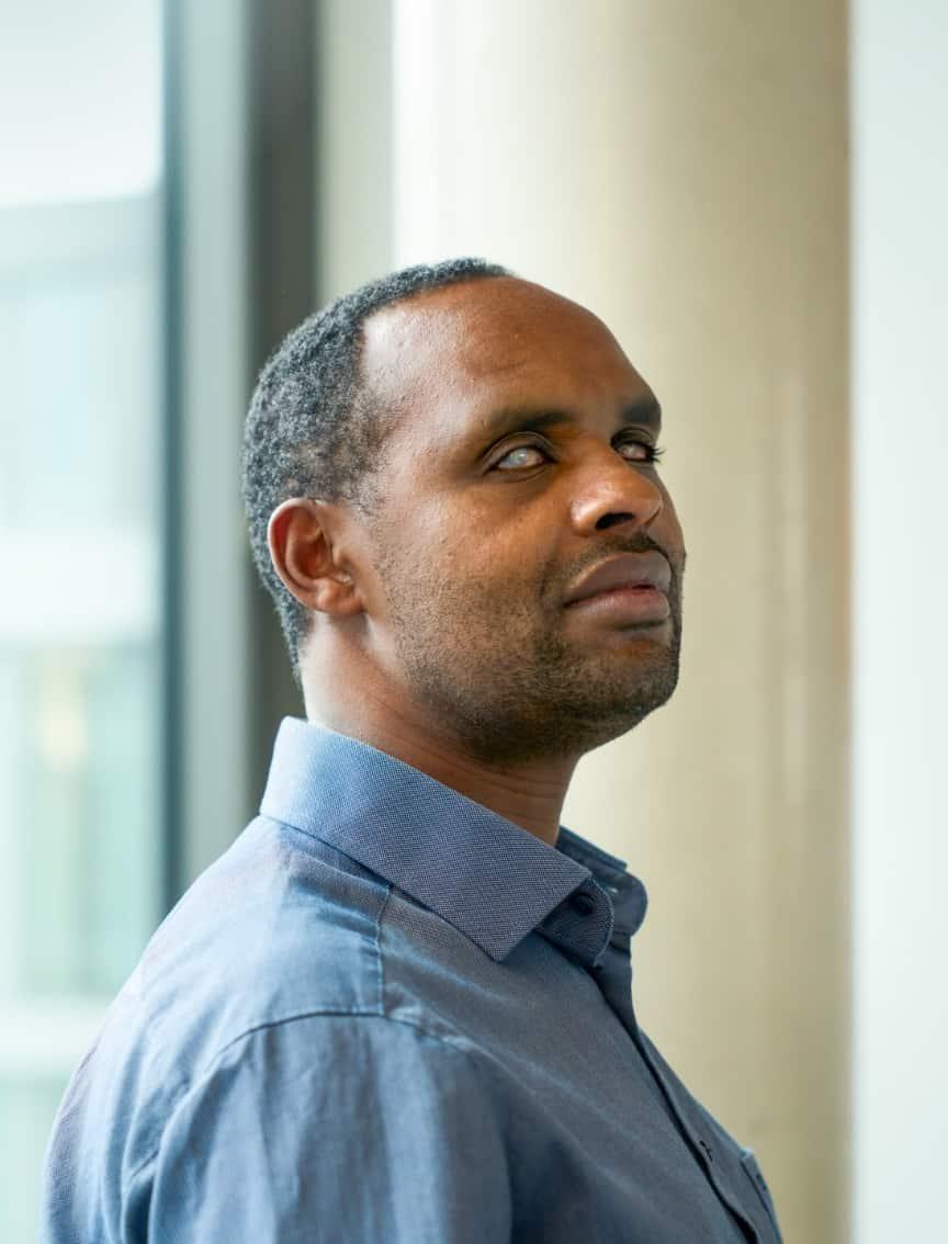 A man in a blue shirt looks to the side, with a large column and a window with daylight in the background.