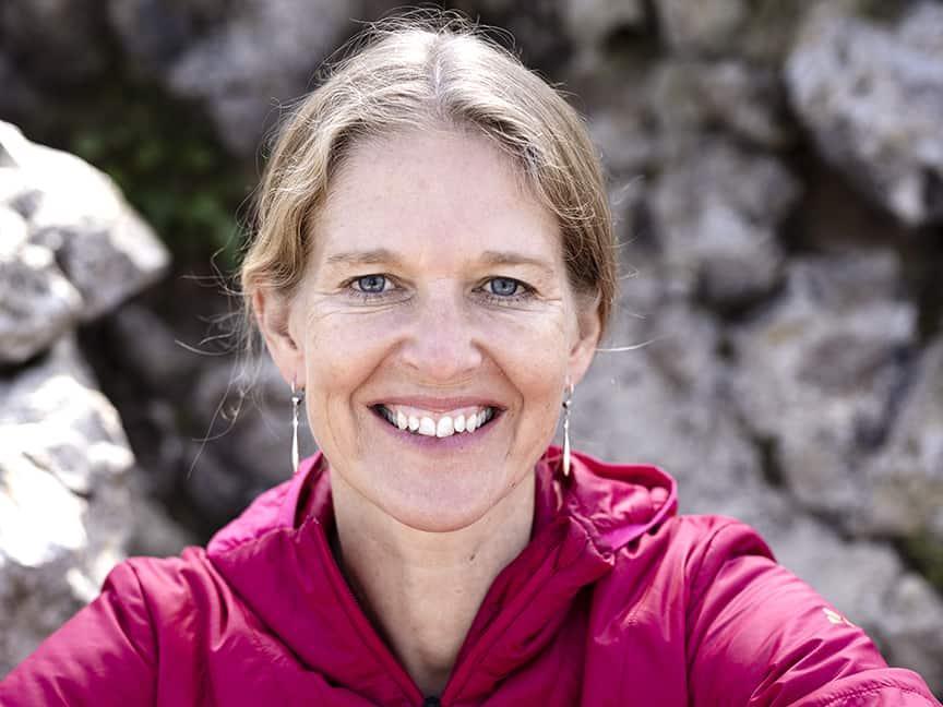 A smiling woman in a red jacket looks directly into the camera, with a rock face in the background.