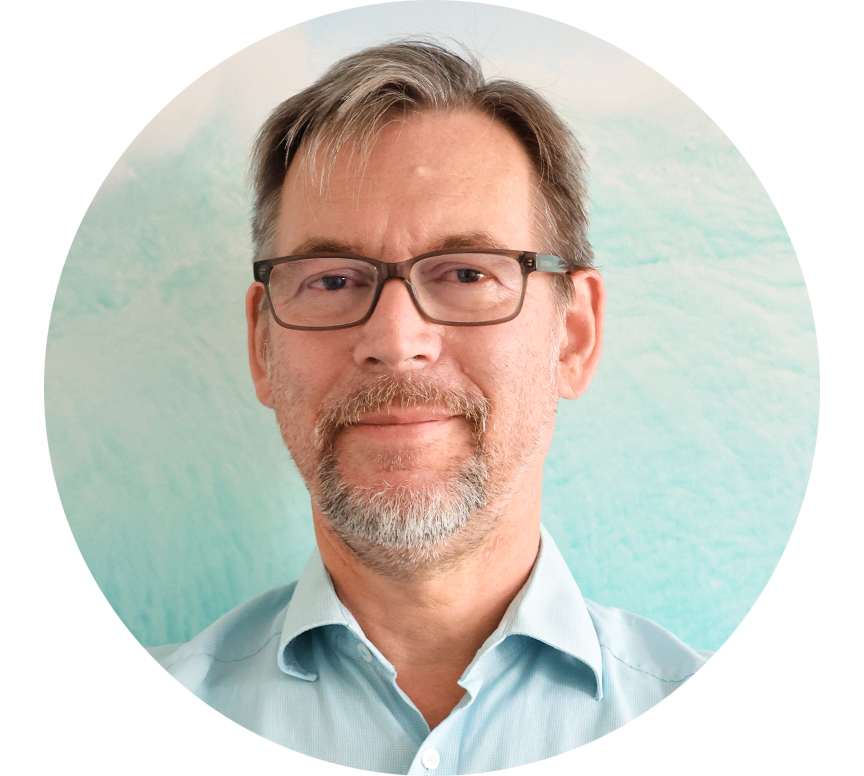 Portrait of an older man with glasses, a grey beard and a light blue shirt in front of a pastel-coloured background.
