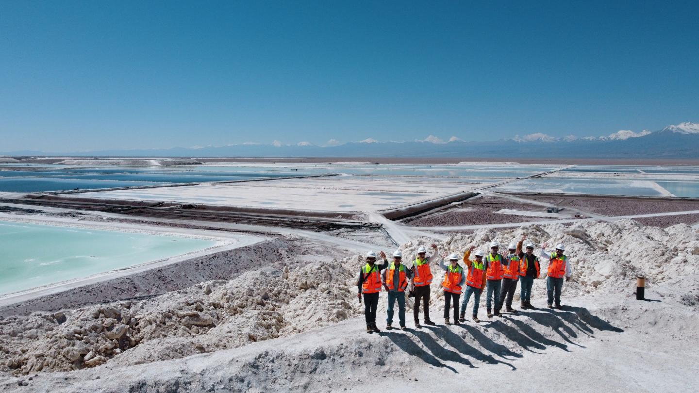 Group of people with safety helmets and bright orange safety vests standing on a hill in a salt mining area with large evaporation ponds; in the background, mountains and a clear blue sky are visible.
