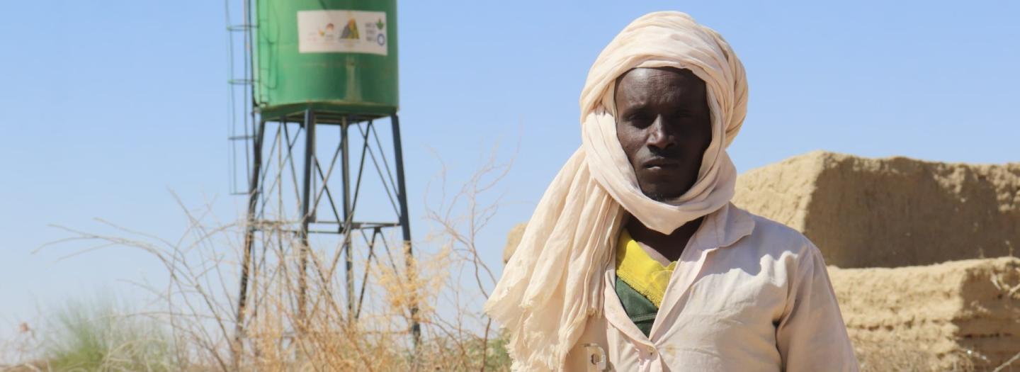 Ein Mann – Ibrahim Yatara – steht in Wüstenkleidung vor einem grünen Wassertank in einer trockenen Landschaft.