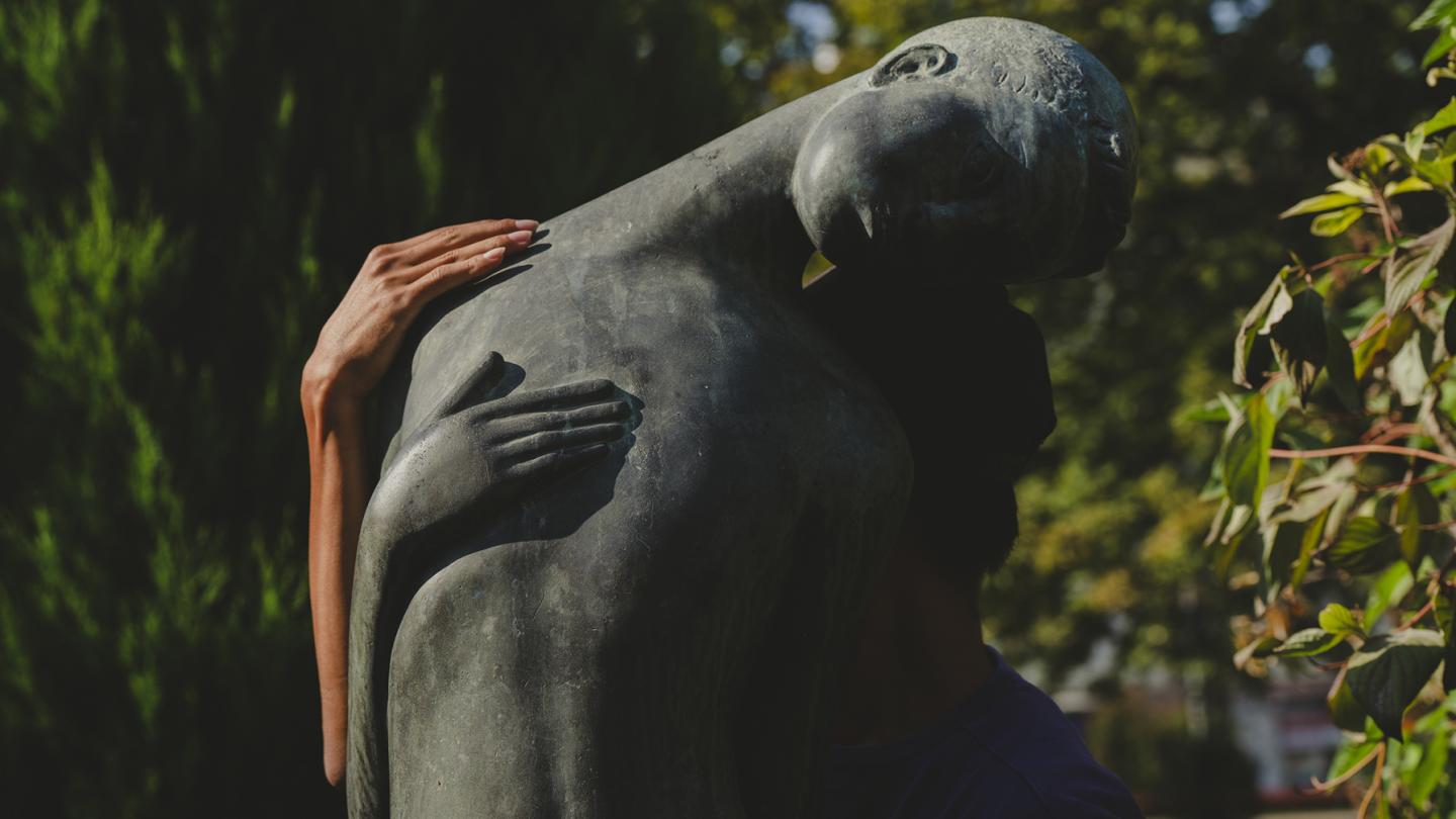 A real person embraces a female bronze statue, with the human and statue hands placed parallel on the back.
