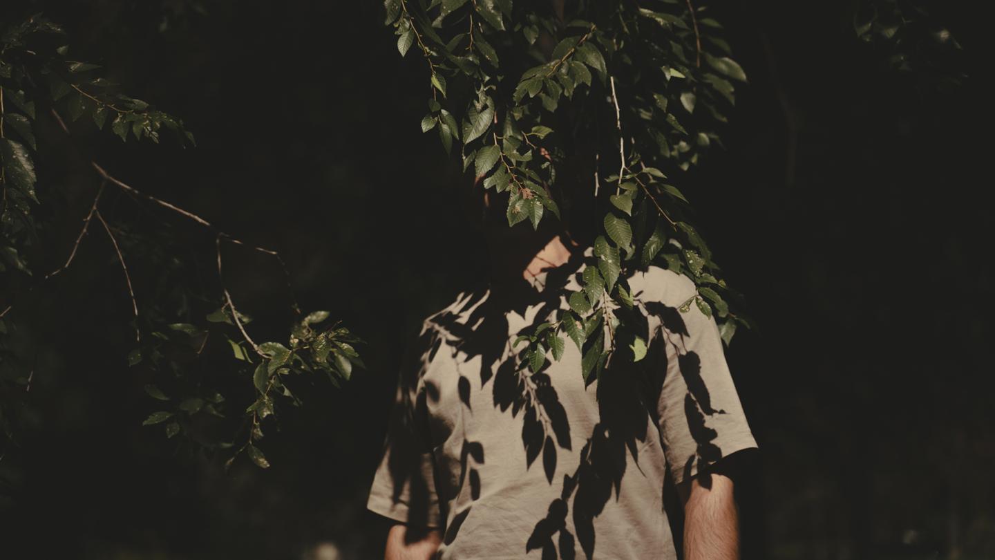 A person stands in the shadow of a tree, their face completely hidden by dense green leaves.