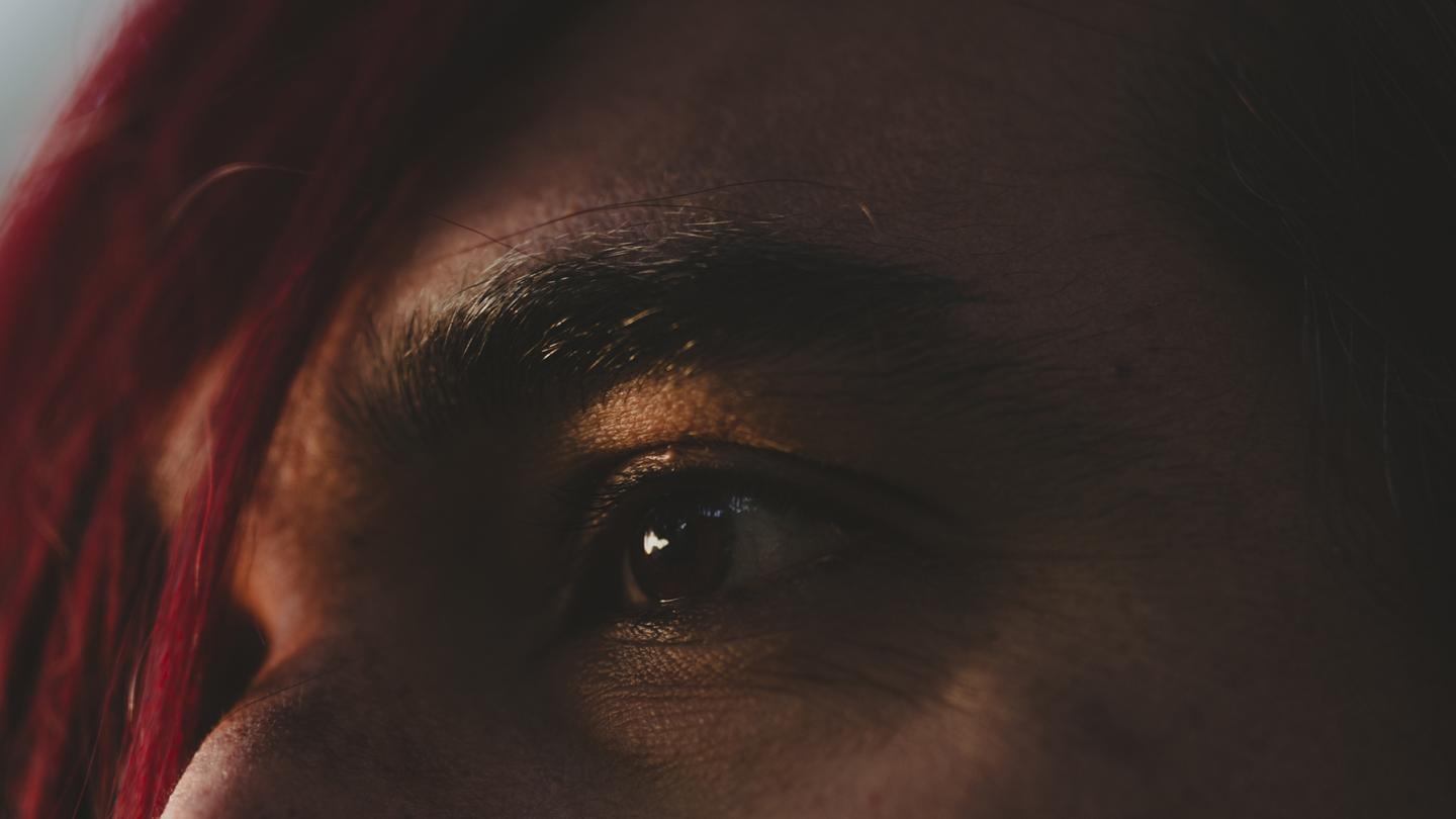 Close-up of a brown eye illuminated by warm light, with reddish hair in the foreground.