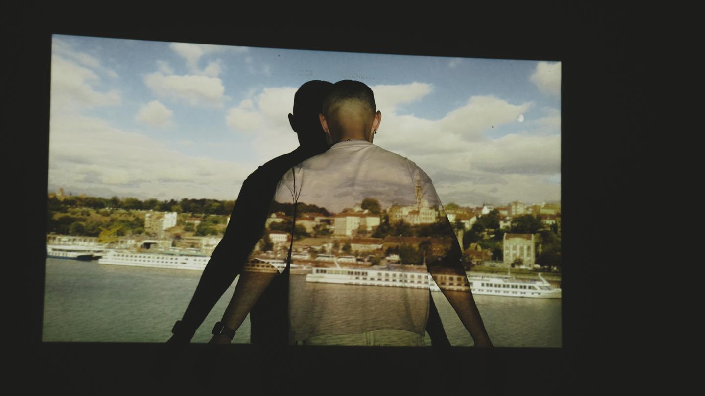 A person stands in front of a projection of a cityscape with a river, with the image cast onto their back.