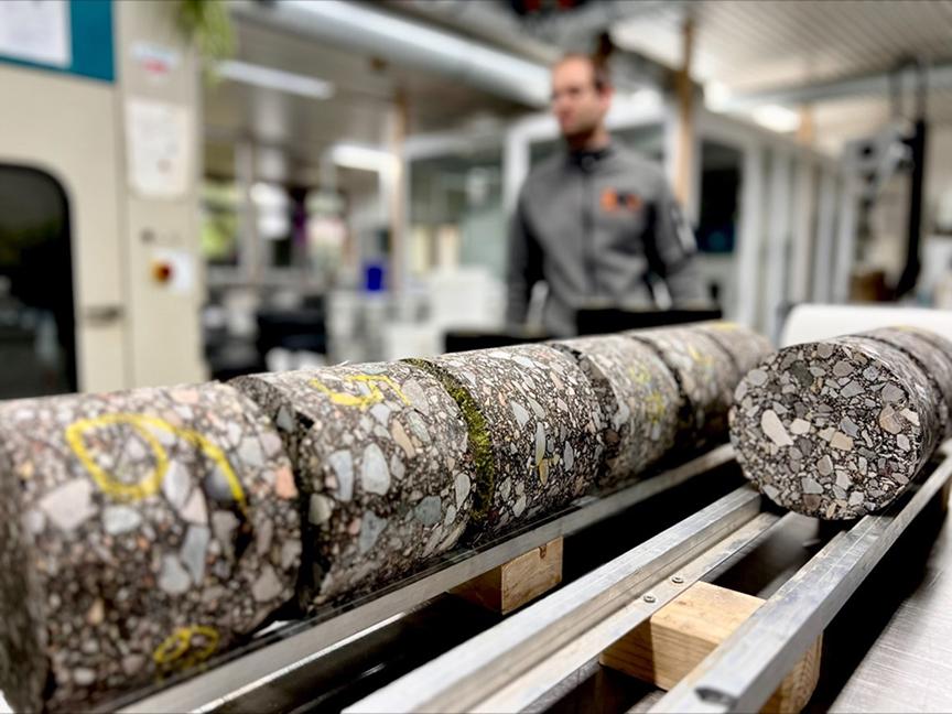 Cylindrical asphalt samples lie in a row on a metal mechanism; in the background is a person in a laboratory.