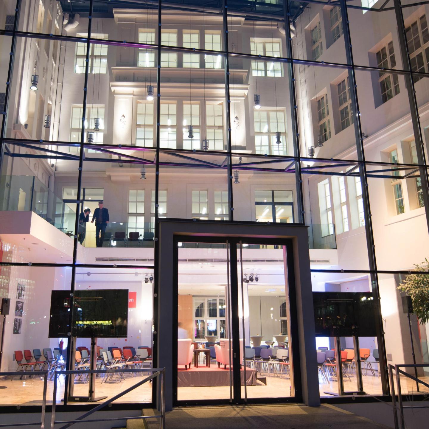 Moderne Glasfassade eines Gebäudes, durch die ein hell erleuchteter Innenhof mit Sitzreihen, Tischen und mehreren Stockwerken sichtbar ist; zwei Personen stehen auf einer Galerie im oberen Bereich.