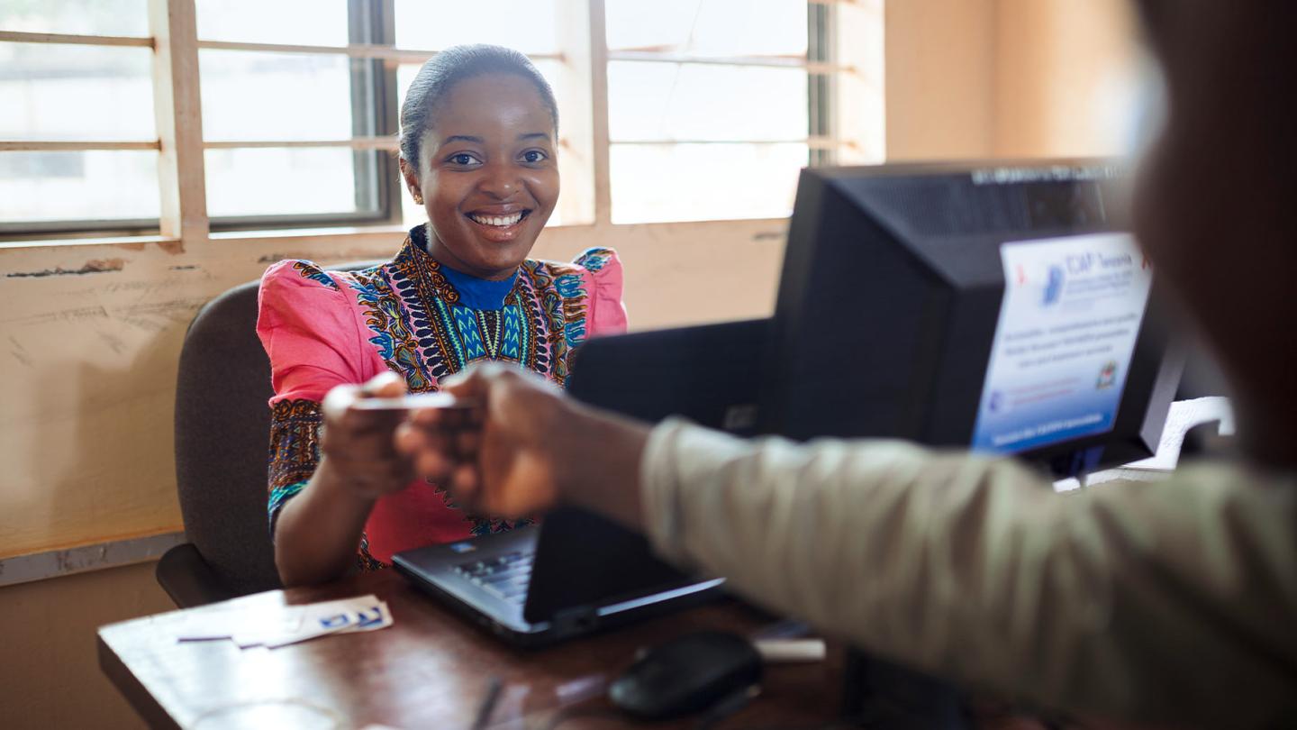 Smiling woman in colorful traditional clothing sits at a desk with a computer and hands a card or document to a person opposite her.
