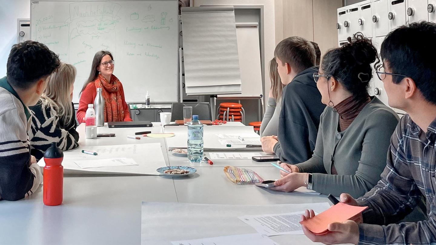 A group of people sitting at a long table in a seminar room while a woman at the end of the table, by the board, speaks; the word ‘sustainability’ is written on the board.