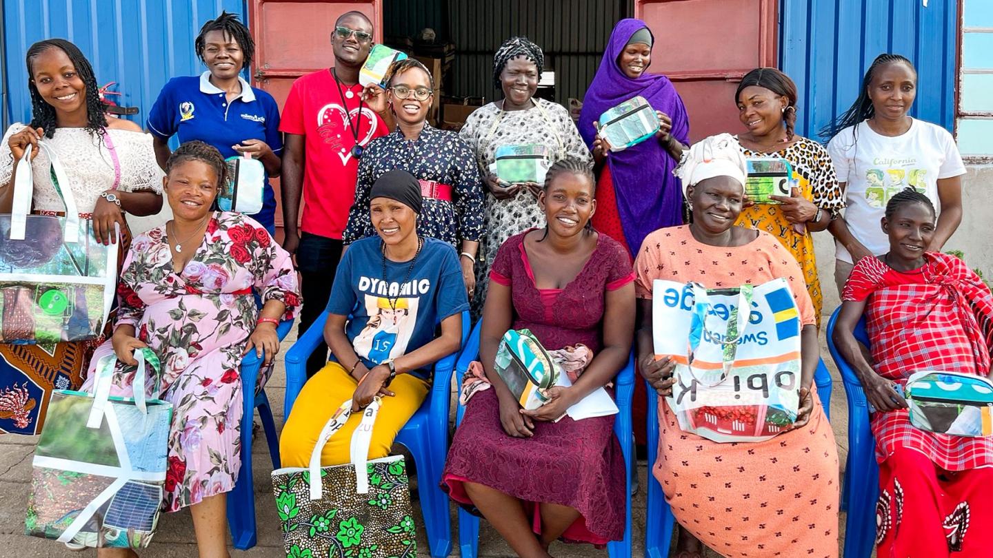 Group photo of women and men in front of a blue building; many are holding bags made of recycled materials that they have sewn together themselves.