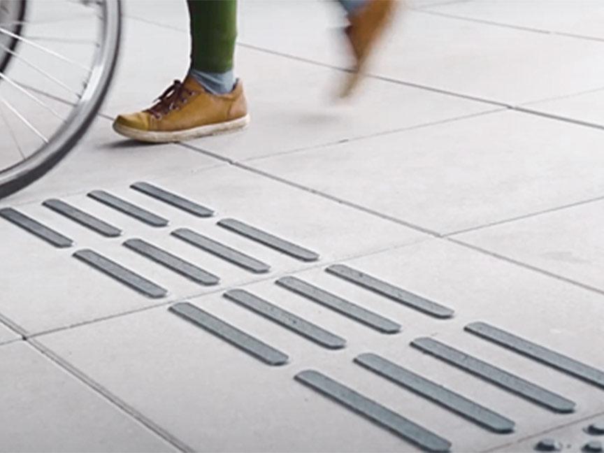 Close-up of a pavement with tactile guide strips for the visually impaired; a wheelchair and a pedestrian are moving over it.