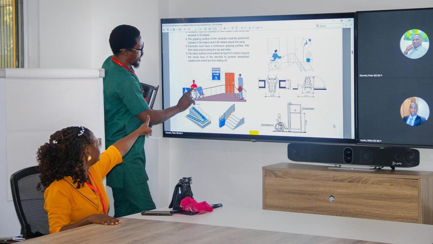 A man and a woman discussing a diagram on accessible infrastructure at a screen while two other participants join the meeting virtually.