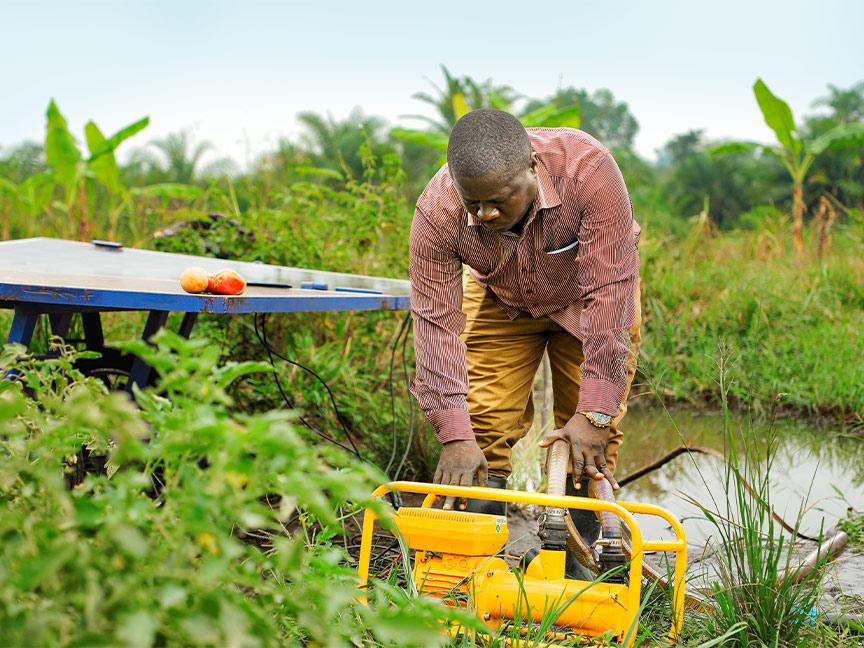 A man working in a field at a yellow water pump; next to it is a table with a solar panel and two tomatoes on it.