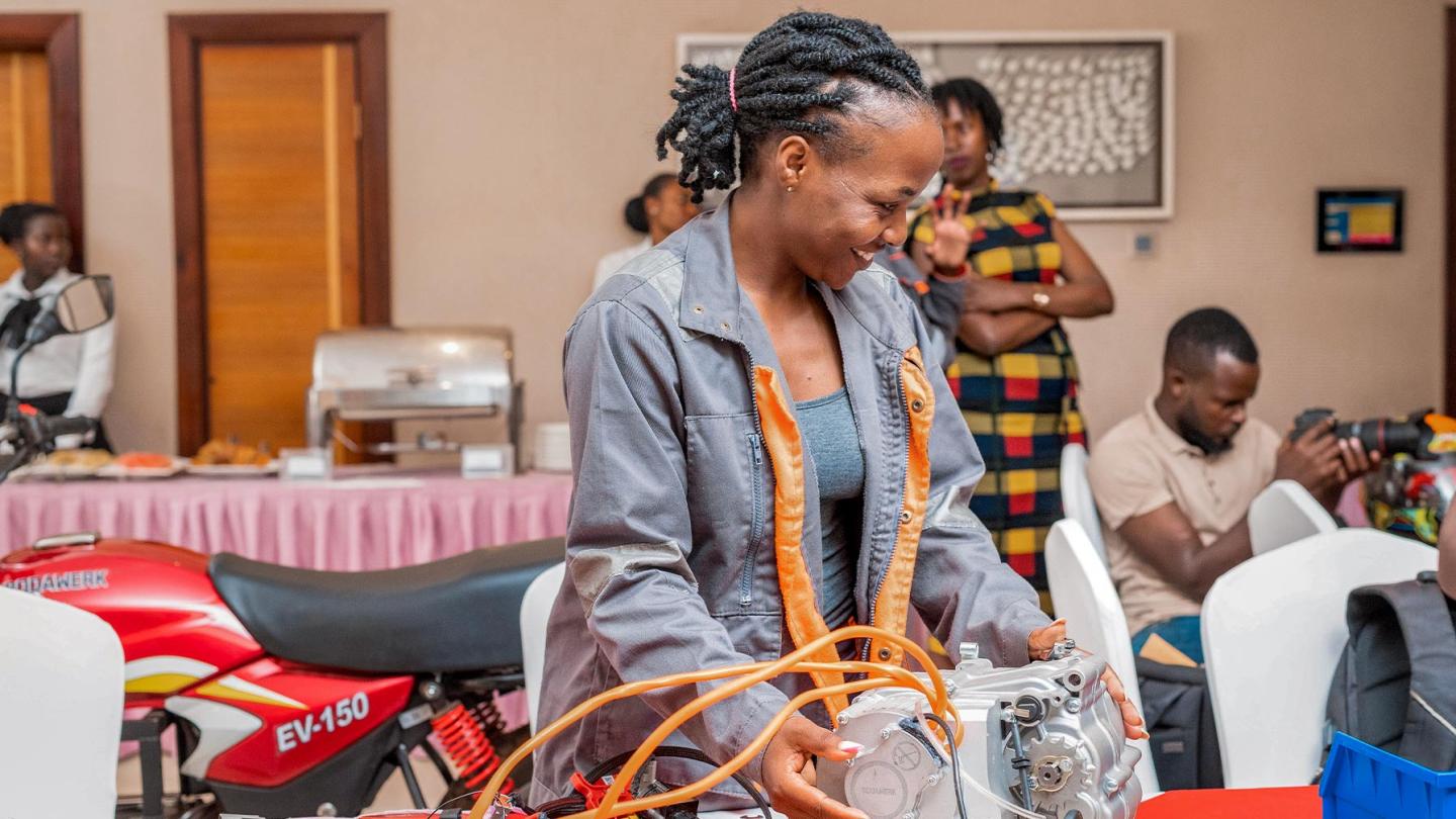 A woman in overalls works on an electric motor; in the background, there is a red motorbike and several people in a function room.
