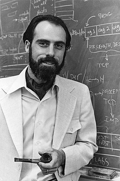 Black-and-white photo of a young bearded man in a light-colored suit holding a pipe, standing in front of a chalkboard with diagrams.