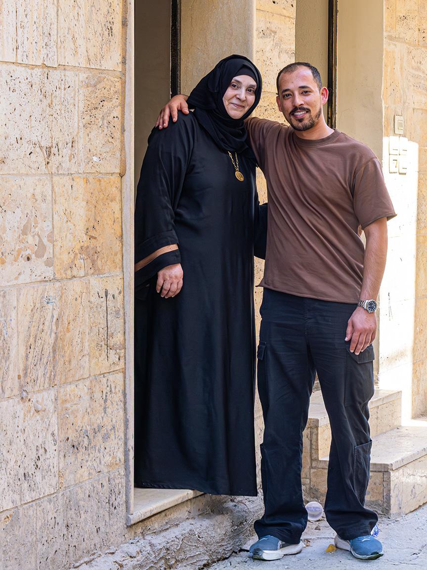 A woman in a black robe and a man in a brown T-shirt pose together in front of a stone facade.