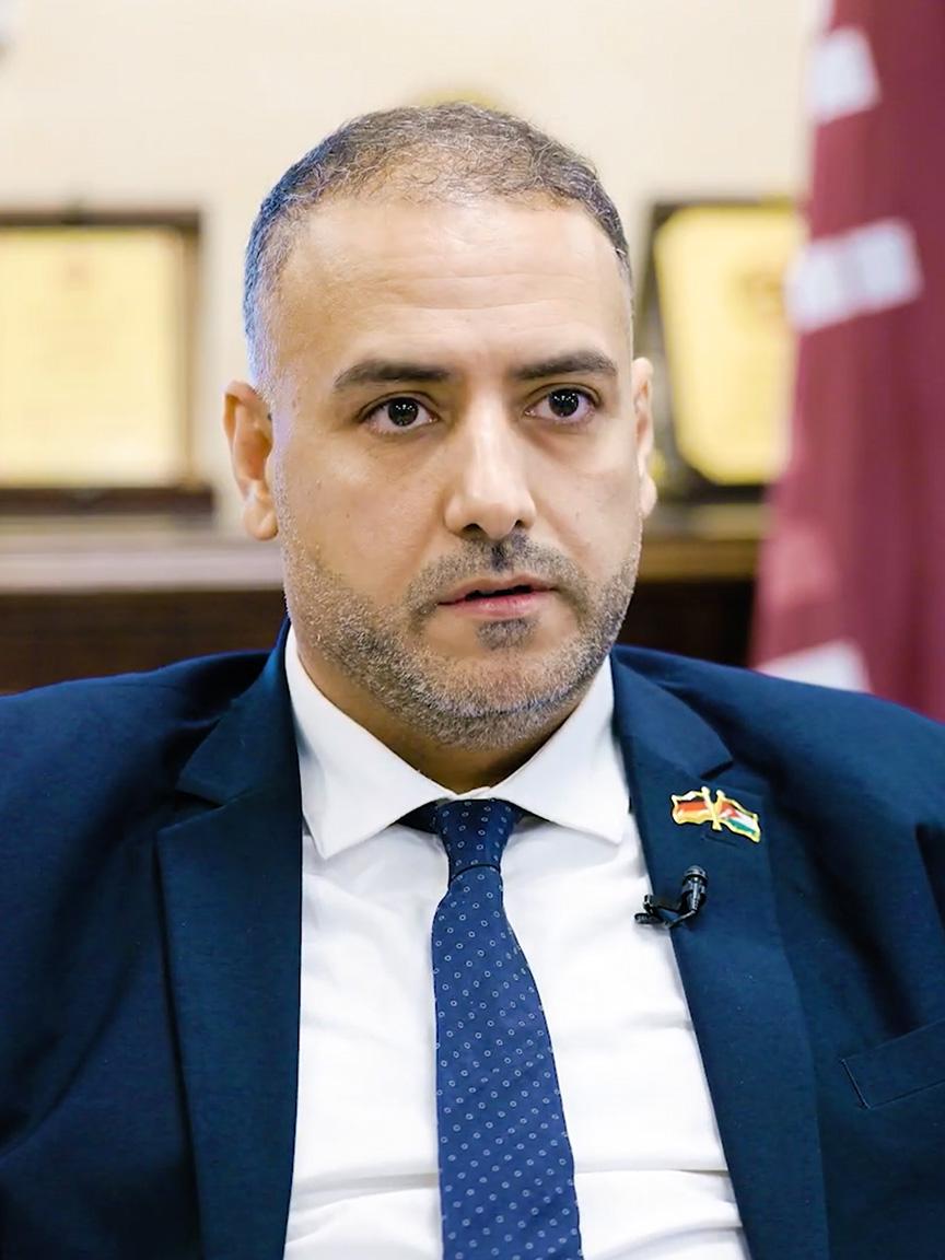 A man in a suit with a microphone and lapel pin speaks in a formal setting.