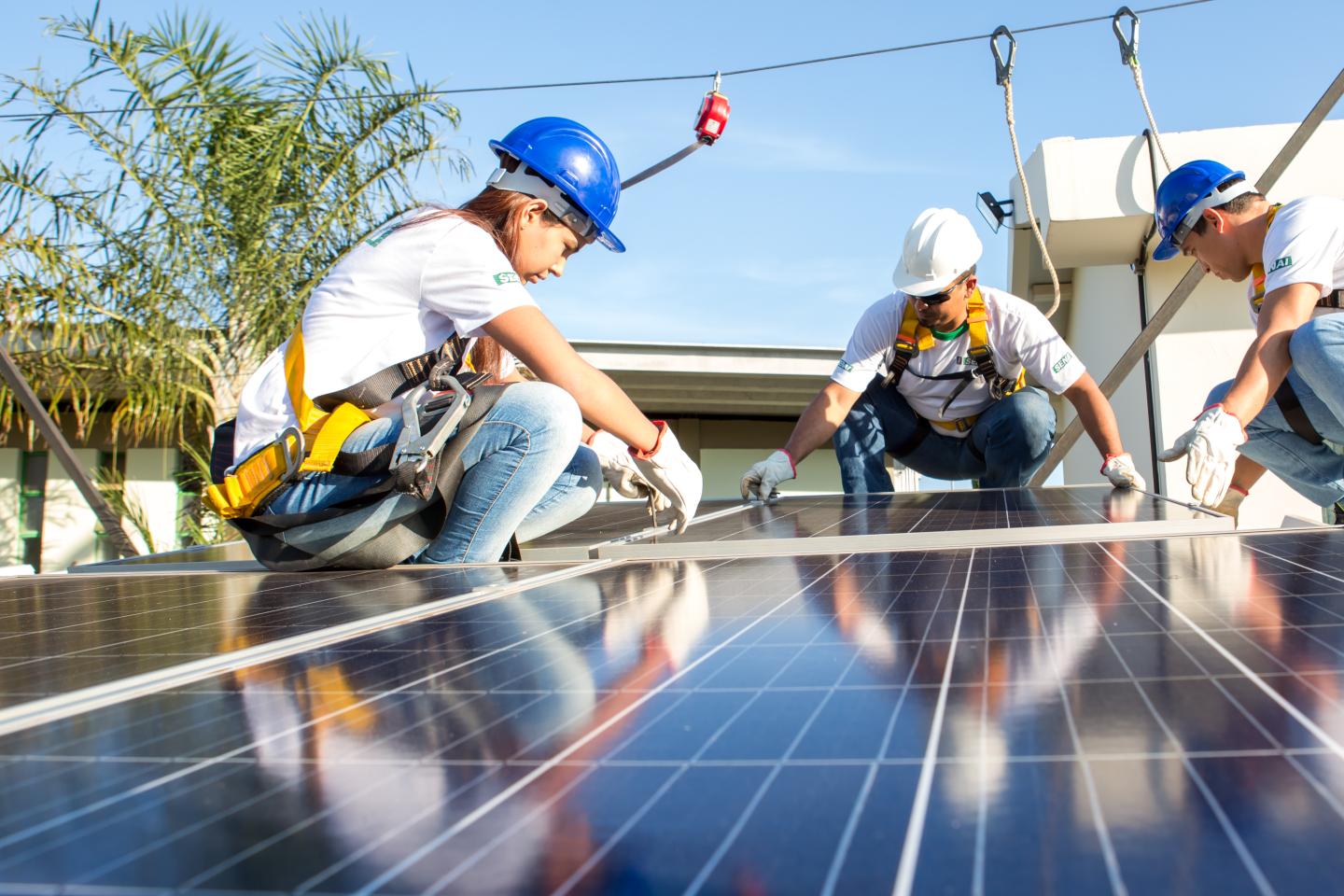 Three people wearing helmets are installing solar panels and are secured by a rope.