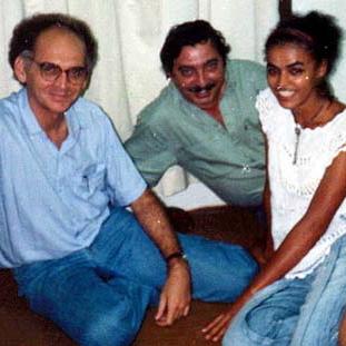 Three people are sitting close together on the floor in front of a white curtain, smiling at the camera.