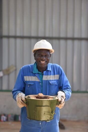 A man in blue overalls and protective clothing is laughing and carrying a cooking stove casing. Copyright: GIZ