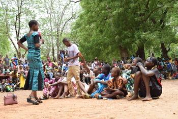 Eine Frau spricht auf einer öffentlichen Veranstaltung zu einer Gruppe sitzender Kinder in Burkina Faso.